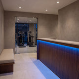 Blue SCOB LED strip lighting beneath a walnut gym reception counter, with seating and exercise equipment beyond; installation illustration.